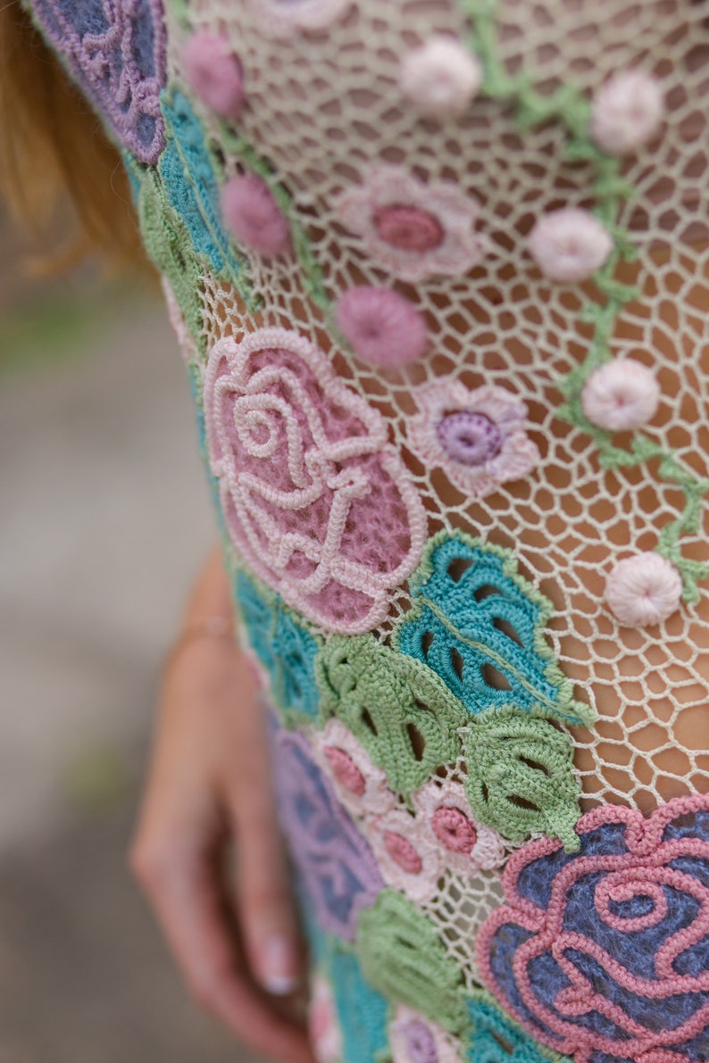 May include: Close-up of a crocheted garment with a floral design. The garment is made of a delicate, white lace with pink, blue, and green flowers. The flowers are intricately crocheted and have a three-dimensional appearance.