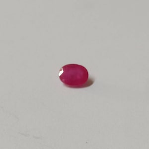 Könnte beinhalten: Ein lebendiger, oval geformter Rubin-Edelstein. Der Edelstein hat eine tiefe, gesättigte rote Farbe mit polierter Oberfläche. Der Edelstein ist vor einem schlichten, hellen Hintergrund platziert.