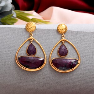 May include: A pair of gold-colored earrings with a hammered texture at the top. Each earring features a teardrop-shaped purple gemstone and a larger, rectangular purple gemstone set within a gold frame. The earrings have a drop design.