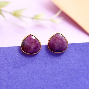 Puede incluir: Un par de pendientes de botón dorados con piedras preciosas rosas oscuras en forma de pera. Las piedras preciosas facetadas están engastadas en un bisel dorado. Los pendientes se muestran sobre una superficie púrpura. Las piedras parecen rubíes.