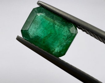 May include: A vibrant green emerald gemstone, rectangular cut, held by silver tweezers against a white background. The gemstone's facets reflect light, showcasing its clarity and color. A close-up view highlights the gem's natural inclusions.