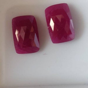 Pode incluir: Duas pedras preciosas vermelhas escuras, facetadas e de formato retangular. As gemas são exibidas em uma superfície branca, mostrando suas superfícies polidas e reflexos internos. As gemas são provavelmente rubis.
