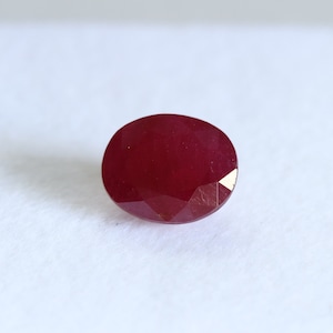 May include: A faceted, oval-shaped ruby gemstone. The gem is a deep, rich red color, with a polished surface that reflects light. The gemstone is set against a white background.