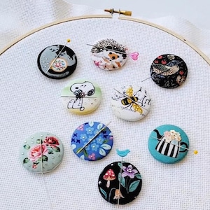 May include: A collection of decorative pin cushions with various designs, including cats, a hedgehog, Snoopy, a bee, floral patterns, and a teapot. Each cushion has a pin inserted, and they are arranged on a white fabric stretched within an embroidery hoop.