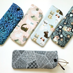 May include: A collection of six fabric eyeglass cases. The cases feature various patterns, including blue water droplets, cats wearing glasses, dogs wearing glasses, and a geometric design. Each case has a snap closure.