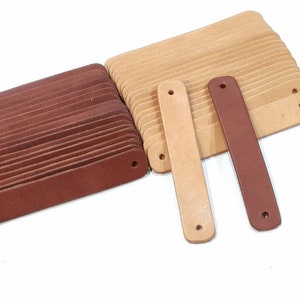 Op de afbeelding: Twee stapels leren stroken, een bruin en een beige, met twee afzonderlijke stroken, een bruin en een beige, liggend op een wit oppervlak. De stroken hebben geperforeerde gaten.