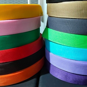 Peut inclure: Une pile de sangles en sangle colorées dans différentes couleurs, notamment jaune, rose, vert, rouge, noir, orange, beige, bleu, violet et gris. Les sangles ont toutes la même largeur et une surface texturée.
