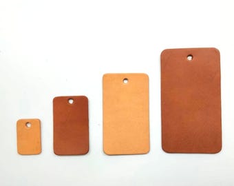 May include: Four leather tags of different sizes, all with a hole punched in the top. The tags are arranged in a row from smallest to largest. The smallest tag is light brown, the next two are a darker brown, and the largest tag is a reddish brown.