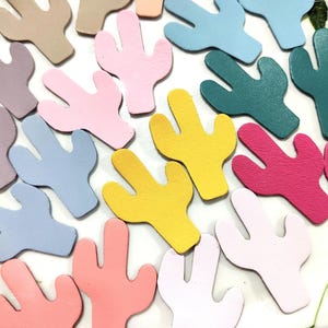 Könnte beinhalten: Eine Sammlung farbenfroher, kaktusförmiger Lederzuschnitte in verschiedenen Farbtönen, darunter Rosa, Gelb, Blau und Grün. Die Zuschnitte sind auf einer Oberfläche mit floralem Muster angeordnet und bilden eine lebendige und verspielte Anordnung. Ideal für Bastelprojekte.
