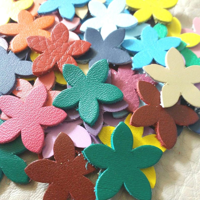 Op de afbeelding: Een verzameling kleurrijke lederen bloemenvormen in verschillende tinten rood, roze, groen, blauw, geel en cr&egrave;me. De bloemen zijn allemaal vijfpuntig en hebben een licht getextureerd oppervlak.