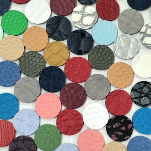 May include: Assortment of small, round leather pieces in various colours and textures. The leather pieces are arranged in a random pattern on a white surface.