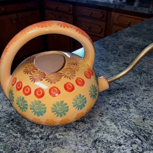 May include: A yellow ceramic watering can with a floral design. The can has a brown handle and a gold metal spout.