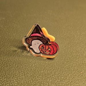 May include: A Halloween-themed pin featuring a ghost wearing a witch hat next to a carved pumpkin. The pin has a clear border and is set against a green background. The witch hat is brown with a pink band.