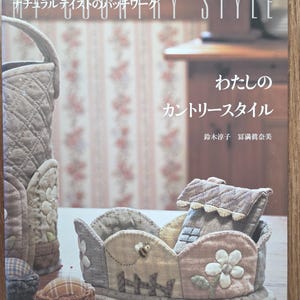 以下が含まれることがあります： 「MY COUNTRY STYLE」と書かれた日本の本。キルトのバスケットや手作りのアイテムが特徴です。表紙には、生地の手工芸品の写真とテキストが掲載されており、カントリースタイルのデザインに焦点を当てています。手作りの布製品に囲まれています。