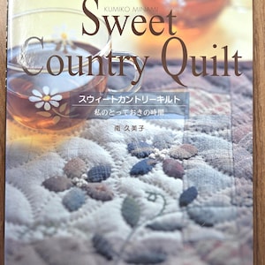 以下が含まれることがあります： 南久美子著「Sweet Country Quilt」というタイトルの本。表紙には、デイジーのデザインの透明なガラスのティーカップと、花の模様のキルト生地のクローズアップが描かれています。日本語のテキストも表示されています。