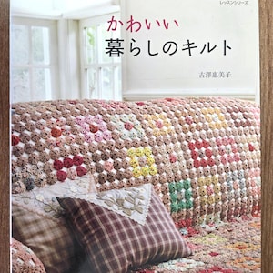 May include: A book titled "かわいい暮らしのキルト" (Kawaii Kurashi no Kiruto) is displayed on a sofa covered with a colorful patchwork quilt. The quilt features a variety of floral and geometric patterns in shades of brown, red, and green. Two plaid pillows are visible.