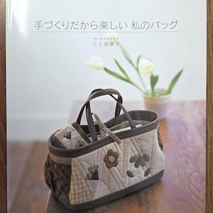 May include: A Japanese language book titled "My Fun Bag Because I Make It", showcasing a handmade quilted bag. The bag features floral and geometric patterns in brown, beige, and grey, with dark brown handles. The book rests on a wooden surface.