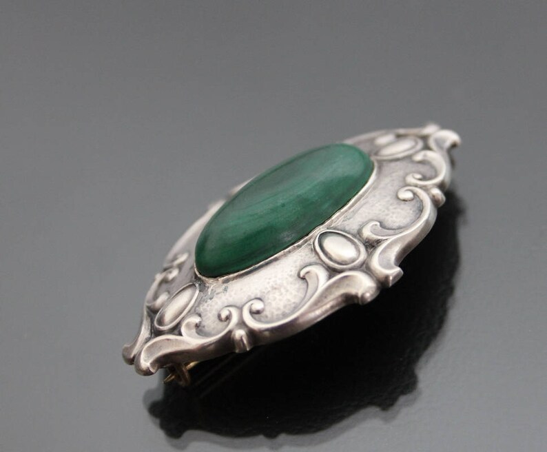 May include: A silver brooch with an oval-shaped green malachite stone set in a detailed, ornate design.