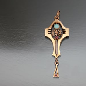 Puede incluir: Un colgante de oro con un diseño en forma de cruz y tres piedras preciosas. El colgante tiene una piedra turquesa en el centro y dos piedras rojas a cada lado. El colgante está suspendido de una cadena de oro.