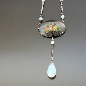 May include: A silver necklace with a large oval pendant featuring a painted floral design. The pendant is surrounded by small, round, opalescent stones. The necklace also has a teardrop-shaped opalescent stone hanging from the bottom.