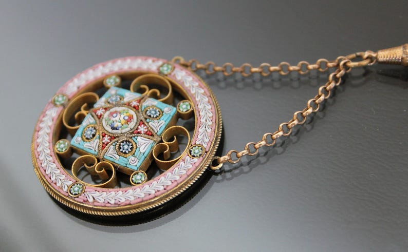 May include: A round pendant necklace with a detailed mosaic design in shades of blue, pink, and white. The pendant is surrounded by a delicate gold-colored frame and hangs from a gold-colored chain.