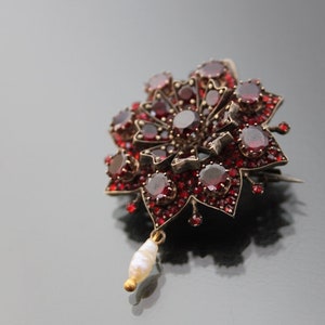 Op de afbeelding: Een zilveren broche met rode granaten en een parelhangertje. De broche heeft de vorm van een bloem met een centrale granaat omringd door kleinere granaten.