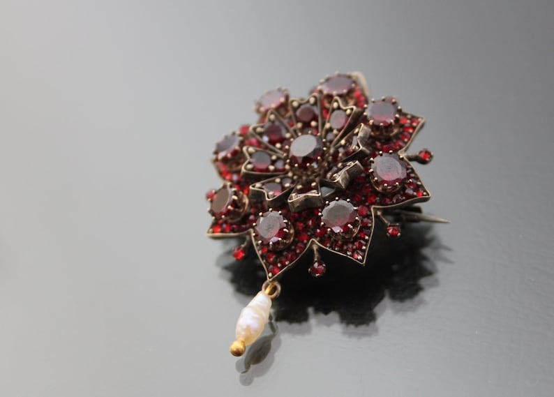 Op de afbeelding: Een zilveren broche met rode granaten en een parelhangertje. De broche heeft de vorm van een bloem met een centrale granaat omringd door kleinere granaten.