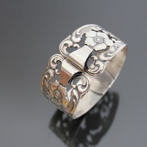 May include: Silver napkin ring with a floral design. The ring is open and has a hinged clasp.