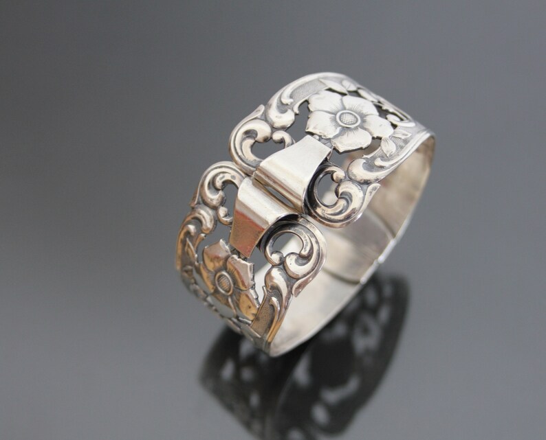 May include: Silver napkin ring with a floral design. The ring is open and has a hinged clasp.