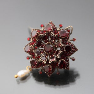 Op de afbeelding: Een zilveren broche met rode granaten en een parel hanger. De broche is gevormd als een bloem met meerdere lagen rode granaten.