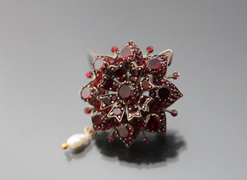 Op de afbeelding: Een zilveren broche met rode granaten en een parel hanger. De broche is gevormd als een bloem met meerdere lagen rode granaten.