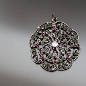 Può includere: Un ciondolo d'argento con un design circolare che presenta un motivo a filigrana e pietre preziose verdi e rosse.