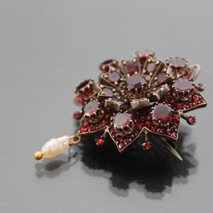 Op de afbeelding: Een zilveren broche met rode granaten en een parel hanger. De broche heeft de vorm van een bloem met een centrale rode granaat omringd door kleinere rode granaten.