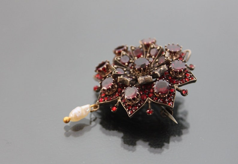 Op de afbeelding: Een zilveren broche met rode granaten en een parel hanger. De broche heeft de vorm van een bloem met een centrale rode granaat omringd door kleinere rode granaten.