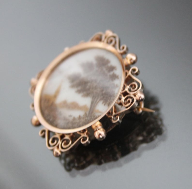 May include: A gold-toned brooch with a delicate, ornate design. The brooch features a round cameo with a white background and a detailed image of a tree in shades of brown.