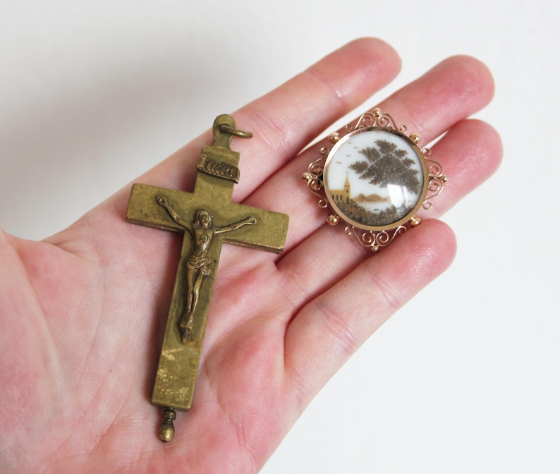 May include: A gold-toned crucifix pendant with a detailed figure of Jesus Christ.  Next to it is a round, gold-toned locket with a glass cover and a small, detailed, hair-art scene inside.