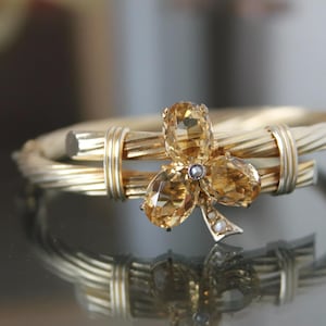 Könnte beinhalten: Ein goldfarbenes Armreif-Armband mit floralem Design. Das Armband hat ein gedrehtes Band und eine dekorative Blume mit drei bernsteinfarbenen Blütenblättern und einem kleinen Perlenakzent. Das Armband spiegelt sich in einer glänzenden Oberfläche.