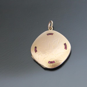 May include: A gold locket pendant with a square shape and a textured surface. The locket is decorated with small red gemstones around the edges.