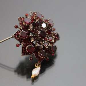 Op de afbeelding: Een zilveren broche met granaat en een parel hanger. De broche heeft de vorm van een bloem met een stervormig ontwerp.