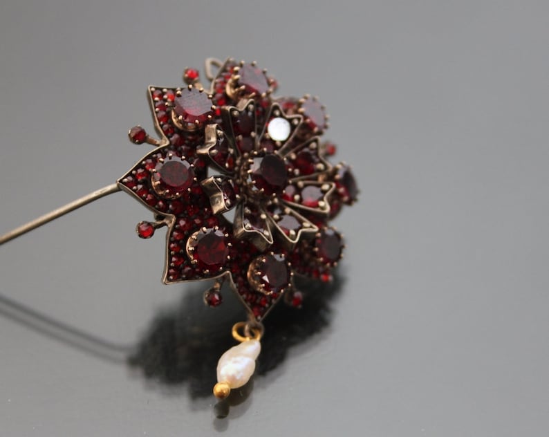Op de afbeelding: Een zilveren broche met granaat en een parel hanger. De broche heeft de vorm van een bloem met een stervormig ontwerp.