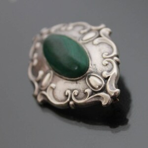 May include: A silver brooch with an oval-shaped green stone set in a decorative frame.