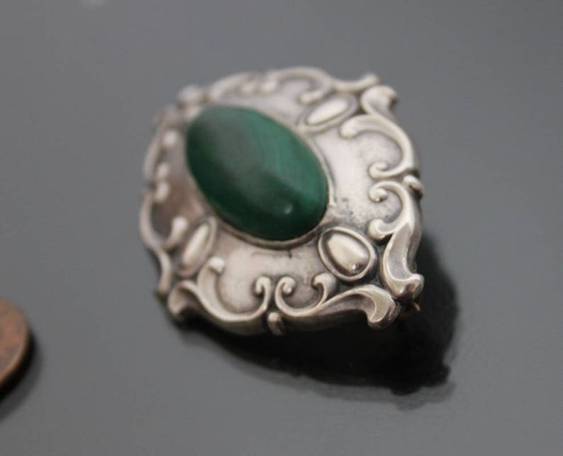 May include: A silver brooch with an oval-shaped green stone set in a decorative frame.