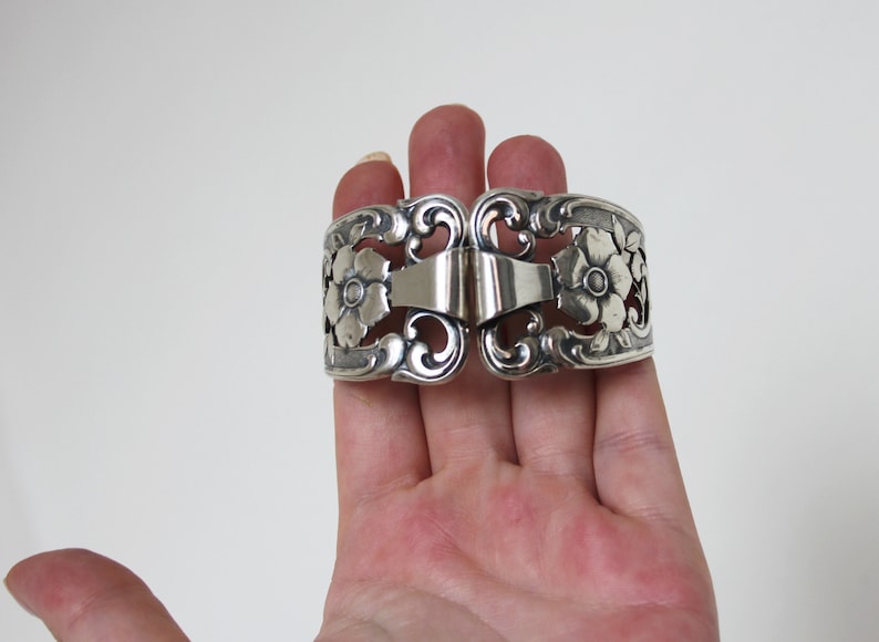 May include: Silver napkin ring with a floral design. The ring is open and has a hinge.