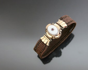 Puede incluir: Un brazalete tejido marrón con un cierre dorado que presenta un camafeo ovalado blanco con una estrella roja en el centro.