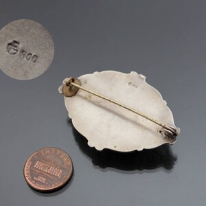 May include: Silver brooch with an oval shape and a detailed design. The back of the brooch has a marking that reads "G 900".