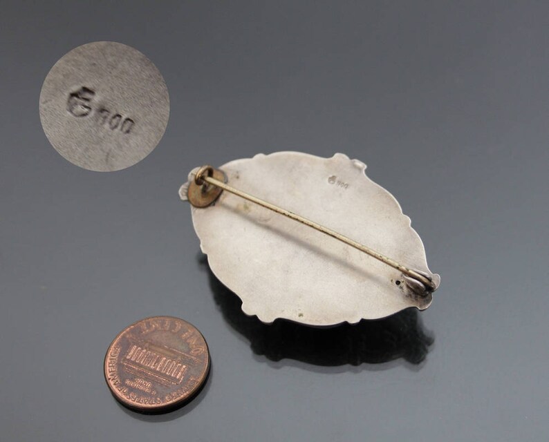 May include: Silver brooch with an oval shape and a detailed design. The back of the brooch has a marking that reads "G 900".