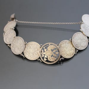 Op de afbeelding: Een zilveren armband met een ketting en meerdere muntvormige bedels. De bedels zijn versierd met ingewikkelde ontwerpen en sommige hebben een gouden afwerking. De middelste bedel heeft een zwarte emaille adelaar.