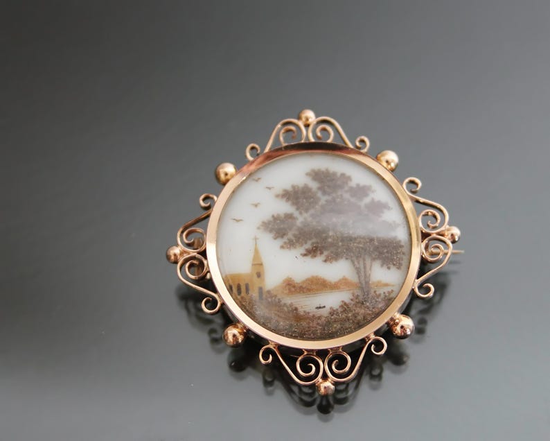 May include: A gold-toned brooch with a round glass cabochon featuring a hand-painted scene of a church and a tree. The scene is made of natural materials like seeds and feathers.
