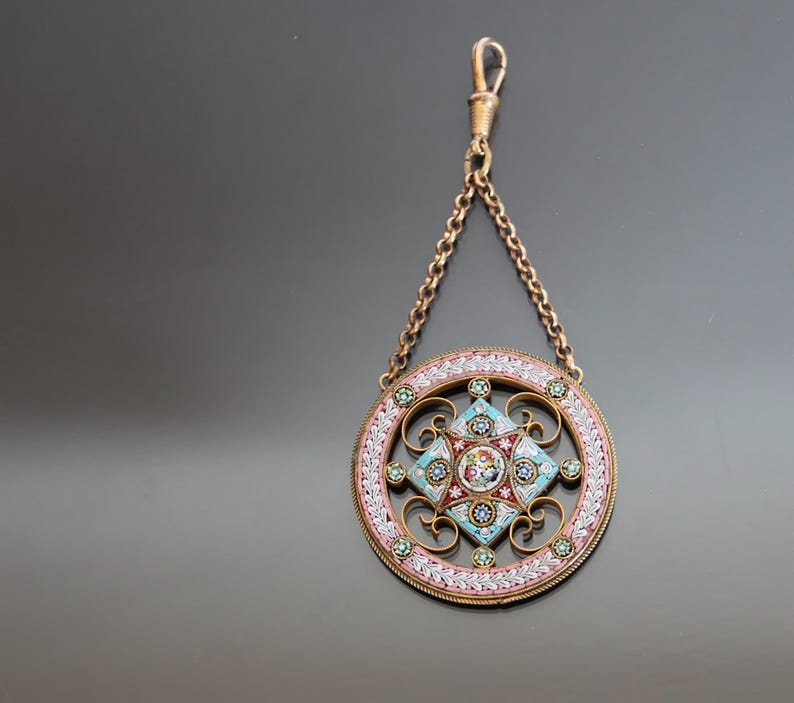 May include: A circular pendant with a delicate floral design in shades of pink, blue, and white. The pendant is made of micro mosaic and is attached to a gold chain.