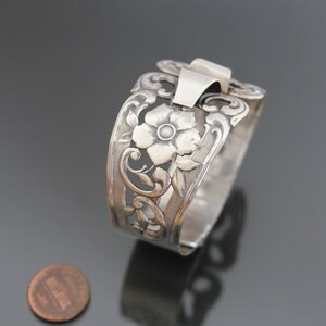 May include: Silver bracelet with a floral design. The bracelet is open and has a clasp on the side. The design features a large flower with a smaller flower on the side.